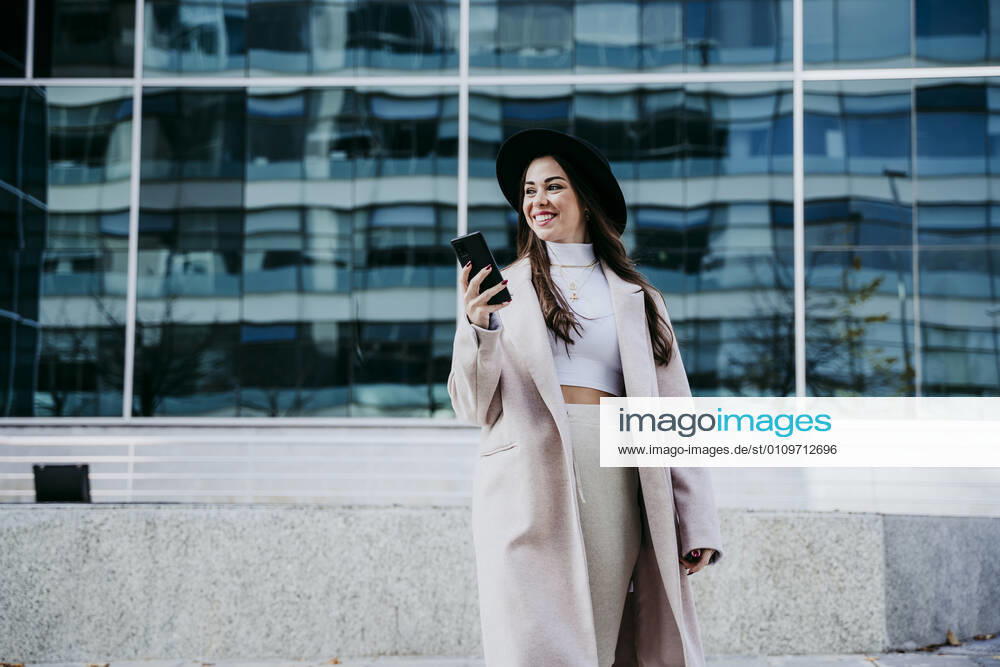Smiling woman with smart phone looking away while standing against building model released Symbolfoto EBBF01484