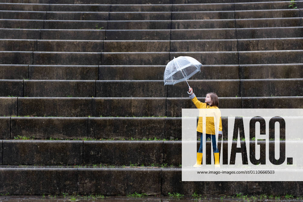 IMAGO Creative: Ende des Sommers Kid in yellow raincoat and rubber boots playing under umbrella along stone steps on rainy day in city