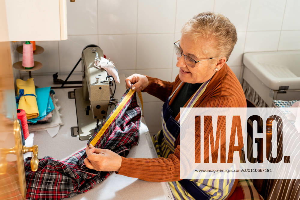 Side view of concentrated senior seamstress sitting at sewing machine using measuring tape on cloth while working in workshop, Model released, Property released