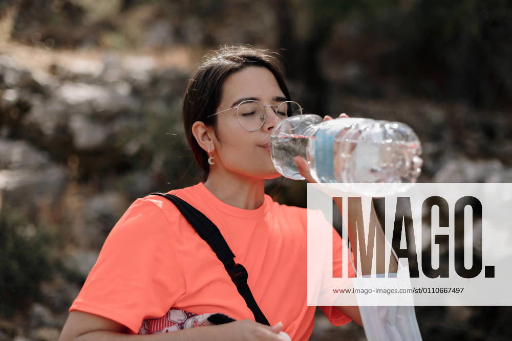 Young woman in casual wear and glasses with medical mask hanging on arm drinking water from plastic bottle while relaxing during hiking in nature