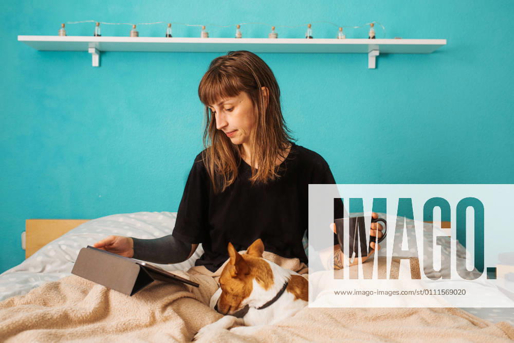 Positive young female in black shirt enjoying hot tea and watching video on tablet while resting on comfortable bed with adorable friendly dog