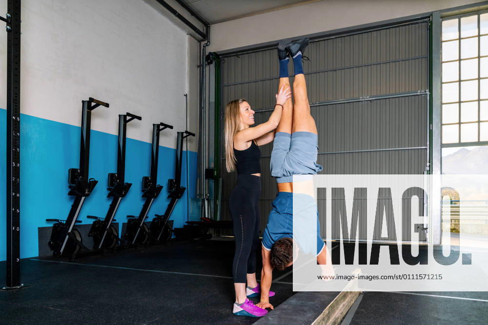 IMAGO Creative: NGO, Rehabilitation und Gesundheitswesen Young smiling female athlete supporting legs of anonymous handicapped sportsman standing on hands during functional training in gym