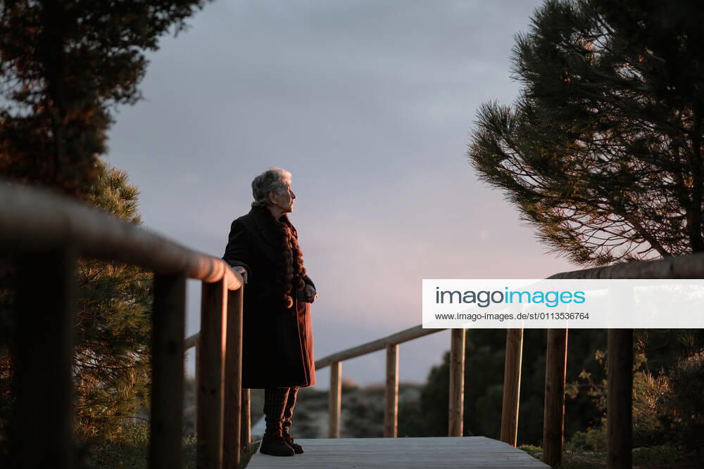 Side view of elderly female traveler in casual clothes standing on wooden pier on sandy beach and enjoying seascape at sunset