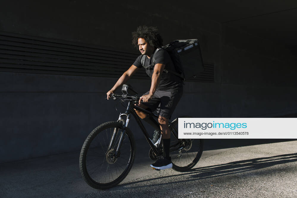 IMAGO Creative, Sommer, Schatten-Pl?tze bei Hitze Side view of ethnic male with delivery box riding bicycle on walkway against wall with shadow in sunlight