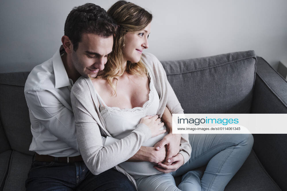 Man with eyes closed embracing pregnant wife while sitting on sofa at home model released Symbolfoto property released EBBF02262