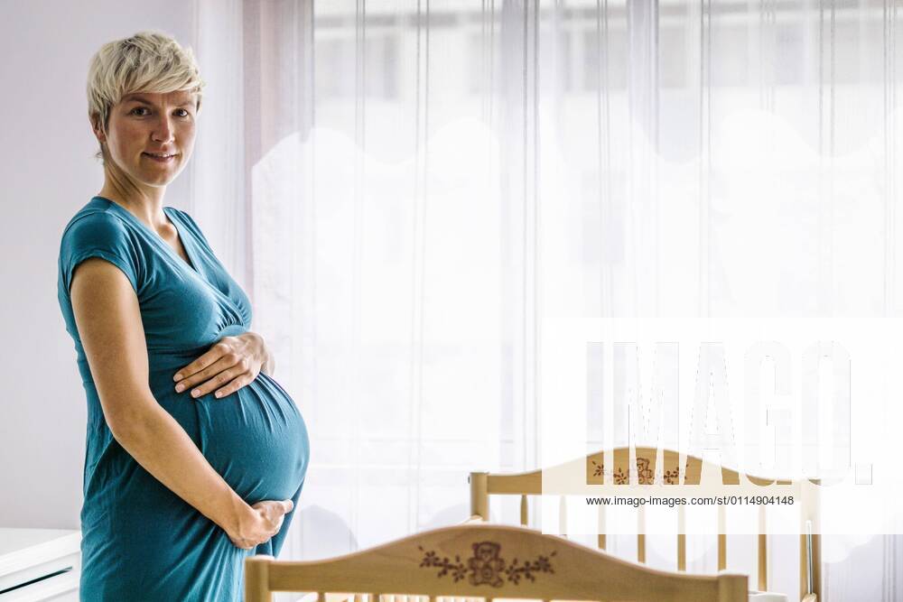 Schwangere Frau hält ihren Bauch durch Babybett neben dem Fenster