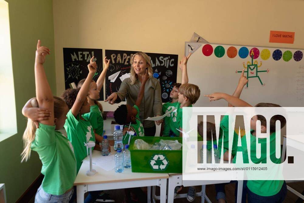 IMAGO Creative: Klimawandel - Umwelterziehung Female school teacher with a group of schoolchildren wearing green t shirts with a white recycling l Front view of a Caucasian female school teacher with long blonde hair and a diverse group of schoolchildren wearing green t shirts with a white recycling logo on them standing around a recycling box, miniature wind turbines and recyclable materials on a table during a lesson in an elementary school classroom, the children raising their hands to answer a question and the teacher pointing, model released, , property released, Cape Town, South Africa, 01.12.2019, 1303839.jpg, people,people,real people,class,classmates,school,elementary school,primary school,classroom,education,lesson,student,pupil,childhood,learn,learning,together,interacting,