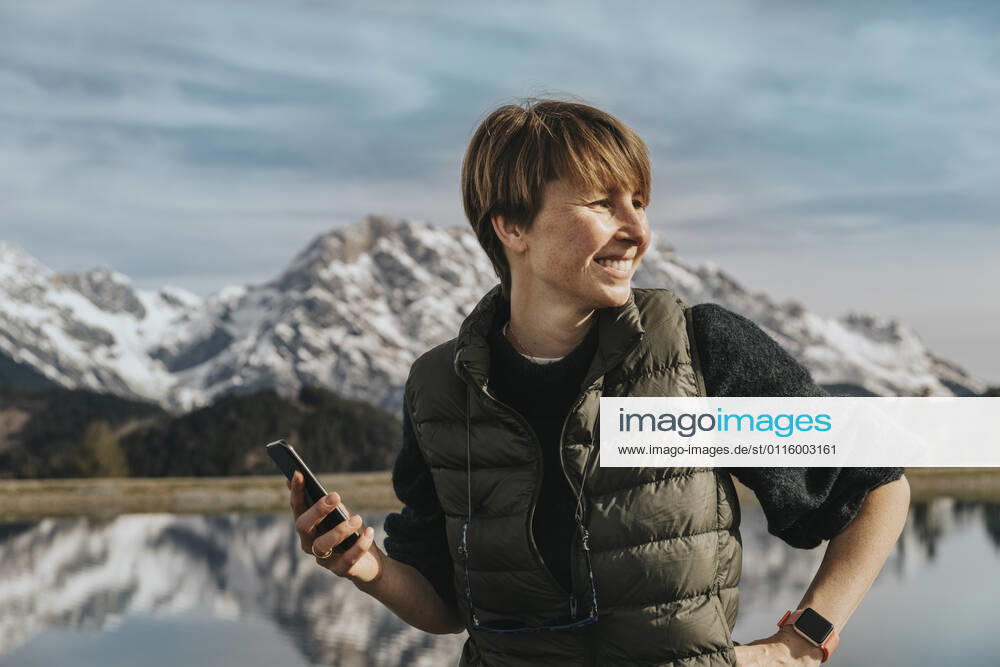 Smiling woman with smart phone looking away while standing at Prinzensee, Hochkoenig massif, Salzburger Land, Austria model released Symbolfoto MFF07221