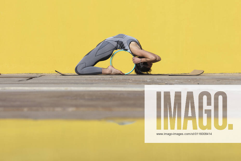 IMAGO Creative: Clickbait, Sport und Fitness Flexible woman with yoga wheel bending over backwards by yellow wall model released Symbolfoto PNAF00668
