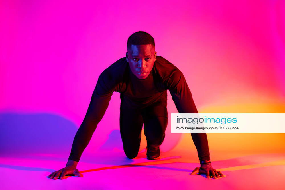 Full length concentrated fit African American male runner standing in crouch start position and looking at camera on colorful background in studio, Model released