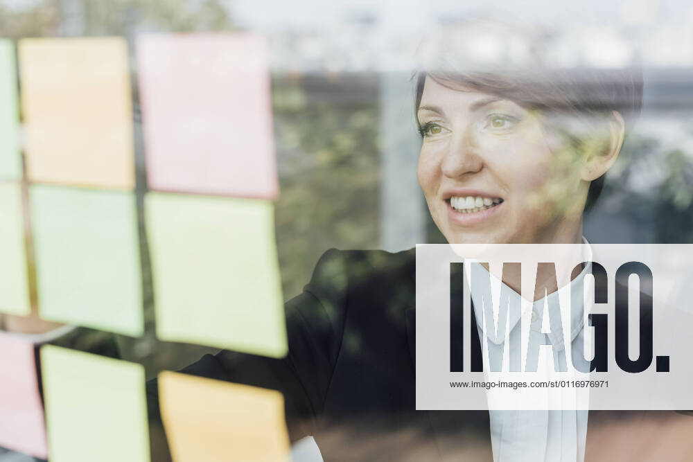 Smiling female entrepreneur looking at adhesive notes on glass wall model released Symbolfoto property released MEUF02264