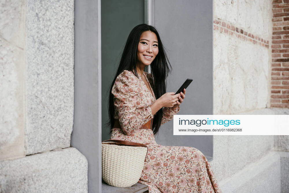 Smiling woman with smart phone looking away while sitting on window sil model released Symbolfoto EBBF03196