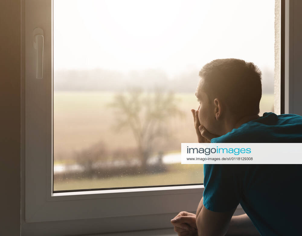 Bavaria, germany - 07 february 2021: young man looks out of the window. he is sad and alone. he supports his head from his hands. the sun shines through the rainy window. lonely and alone concept.