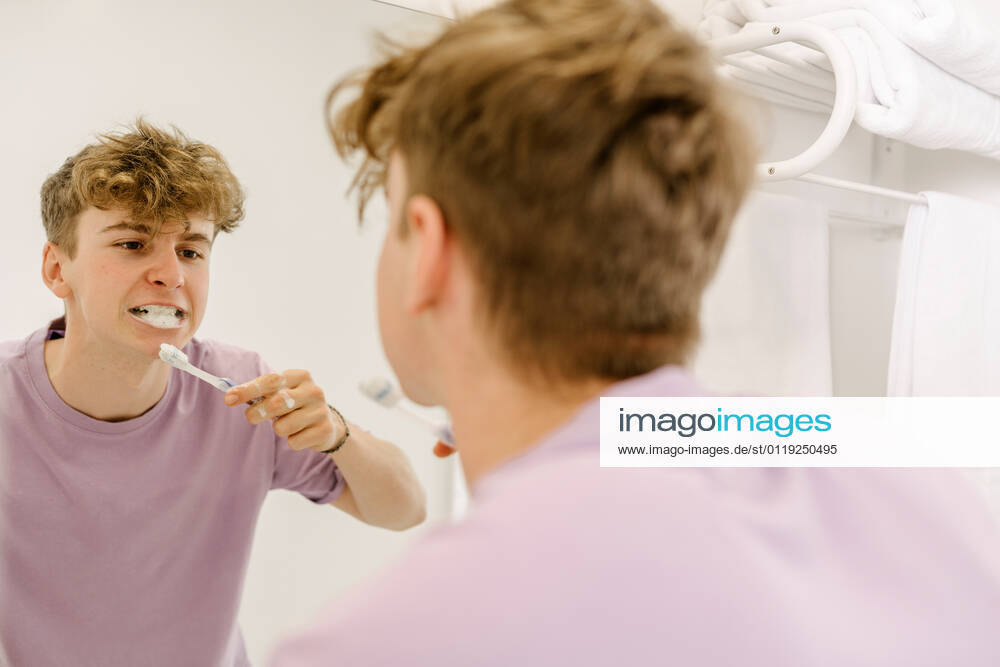 Back view of concentrated male teenager with ginger hair in t shirt brushing teeth using toothpaste and looking in mirror in bathroom, Model released