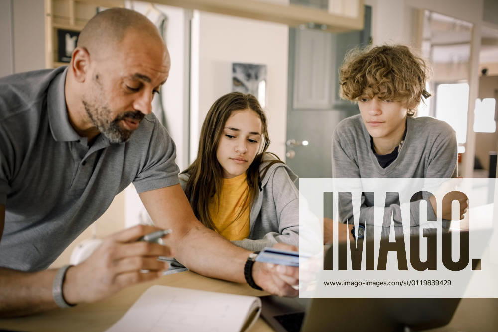 Brother and sister looking at father explaining finance with credit card at kitchen island Sweden, Stockholm, model released, property released