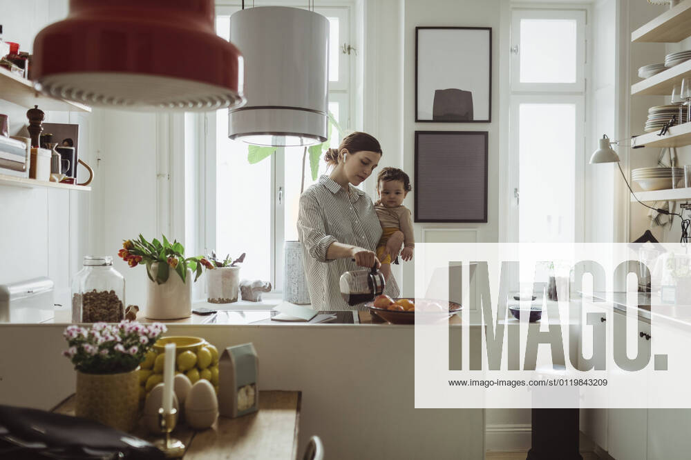 IMAGO Creative, Internationaler Frauenmonat, Entrepreneurinnen Female professional pouring coffee while carrying toddler son in kitchen Sweden, Stockholm, model released, property released