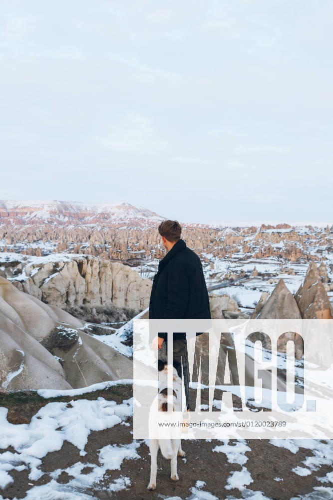 Back view of unrecognizable man in warm clothes with loyal dog in snowy hill against misty mountains in overcast weather in Turkey, Model released
