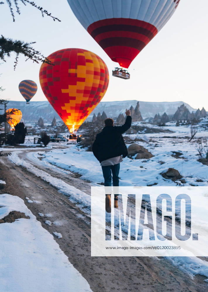 Back view of unrecognizable man in warm clothes with arm raised running to vivid colored air balloon flying up into gray sky over foggy snowy mountain ridge in Turkey, Model released