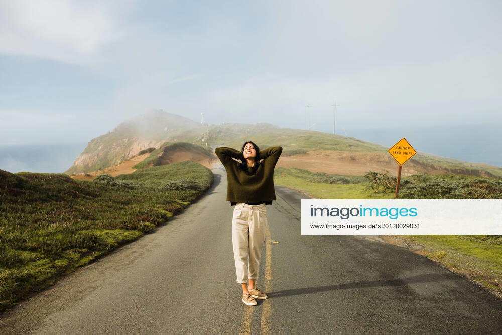 Full length of cheerful woman holding hands behind head in enjoyment of sunlight standing on empty route of coastal cliff in California, Model released