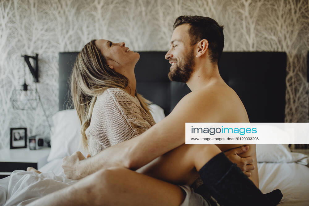 Cheerful young man and woman smiling and cuddling while sitting on comfortable bed at home together, Model released