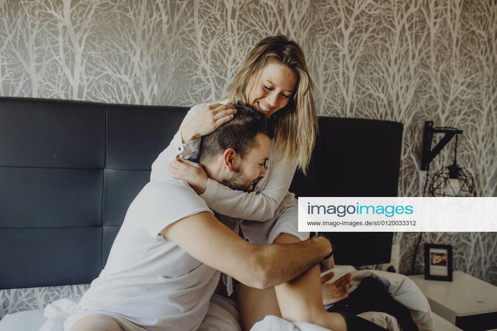Cheerful young man and woman smiling and cuddling while sitting on comfortable bed at home together, Model released