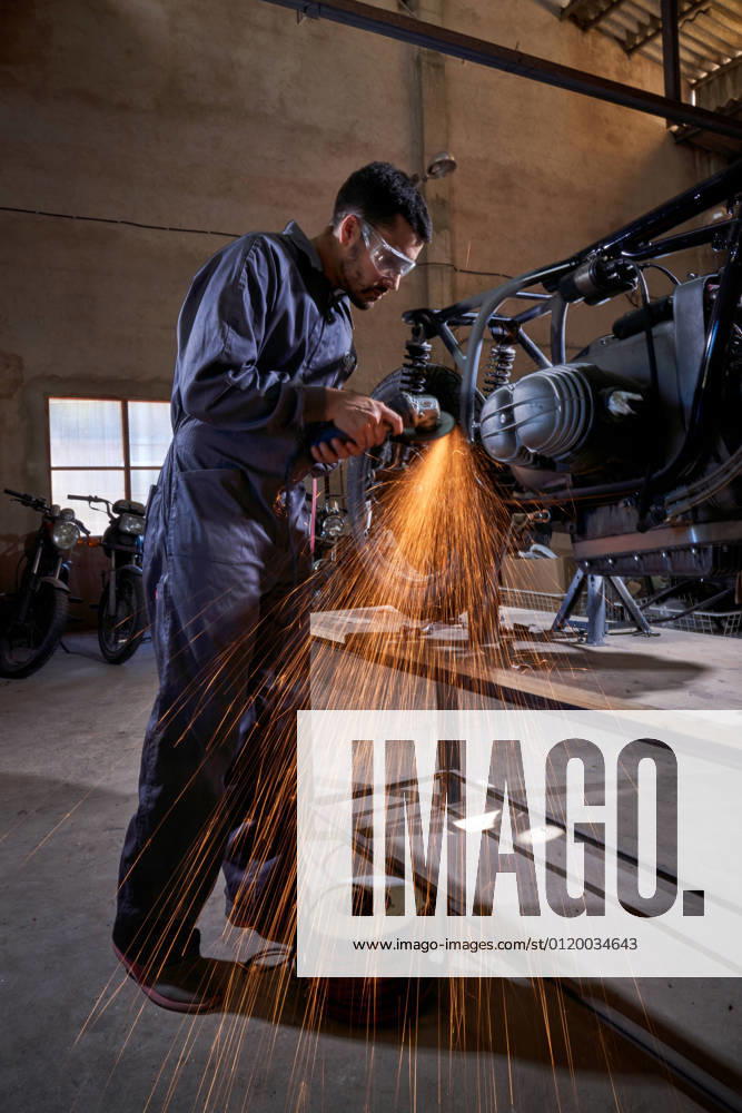 Bearded worker in overall using angle grinder while polishing metal details of custom motorcycle in workshop