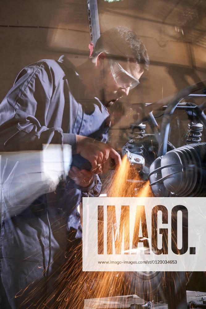 Bearded worker in overall using angle grinder while polishing metal details of custom motorcycle in workshop