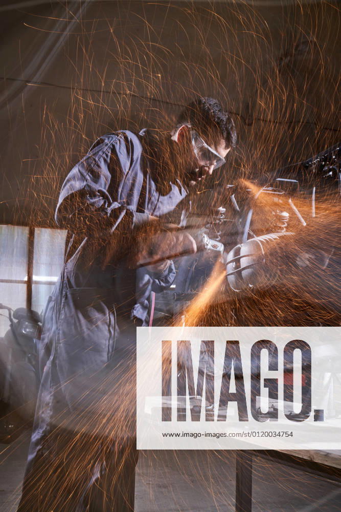 Bearded worker in overall using angle grinder while polishing metal details of custom motorcycle in workshop