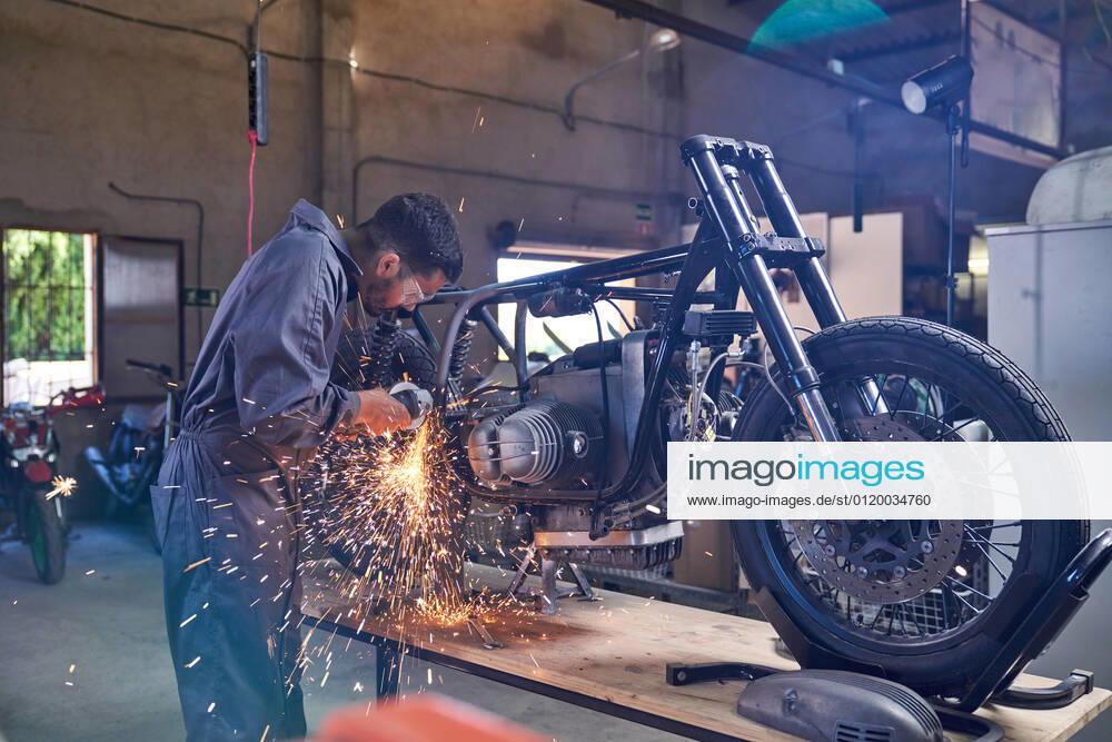 Bearded worker in overall using angle grinder while polishing metal details of custom motorcycle in workshop