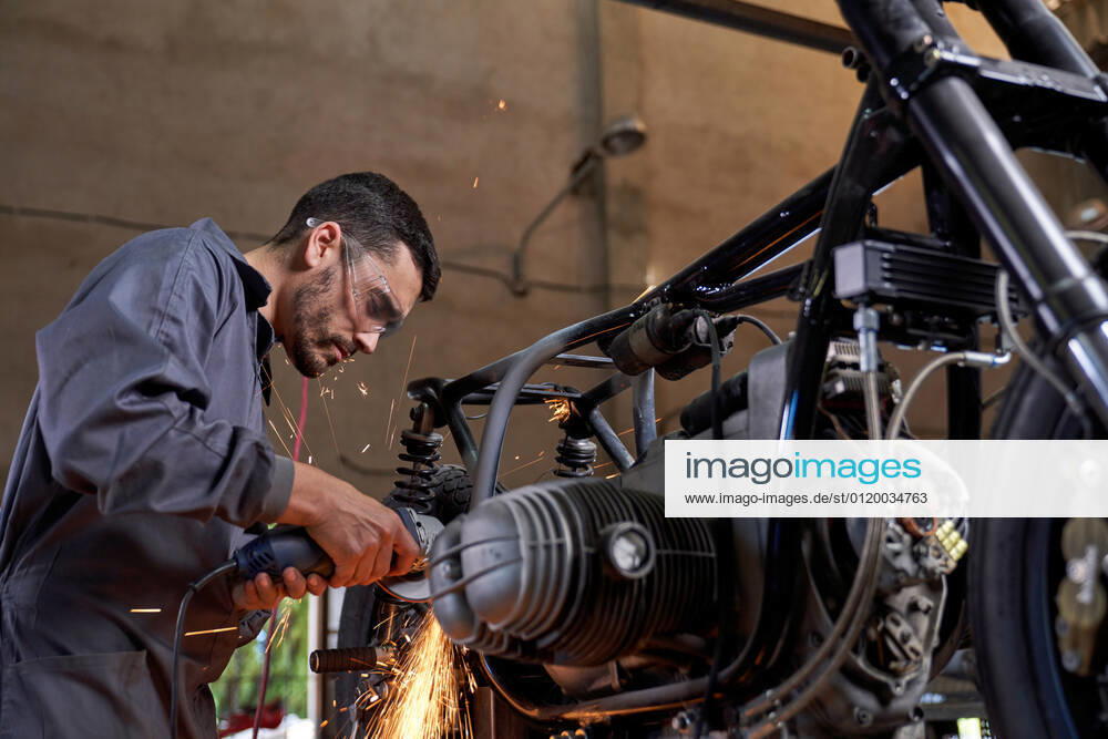 Bearded worker in overall using angle grinder while polishing metal details of custom motorcycle in workshop