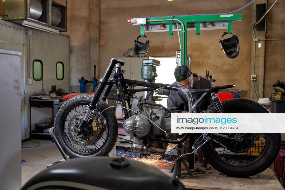 Bearded worker in overall using angle grinder while polishing metal details of custom motorcycle in workshop