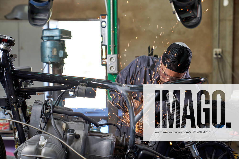 Bearded worker in overall using angle grinder while polishing metal details of custom motorcycle in workshop