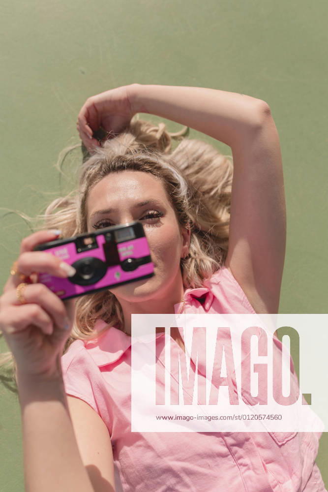 IMAGO Creative, Lifestyle, Barbiecore From above young glad female in pink dress taking pictures on instant photo camera while lying on ground on sunny day