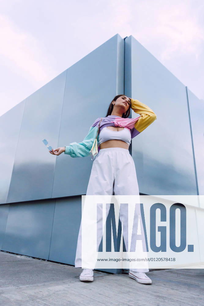 Confident female wearing jeans with top and colorful shirt with stylish sunglasses standing near wall, Model released
