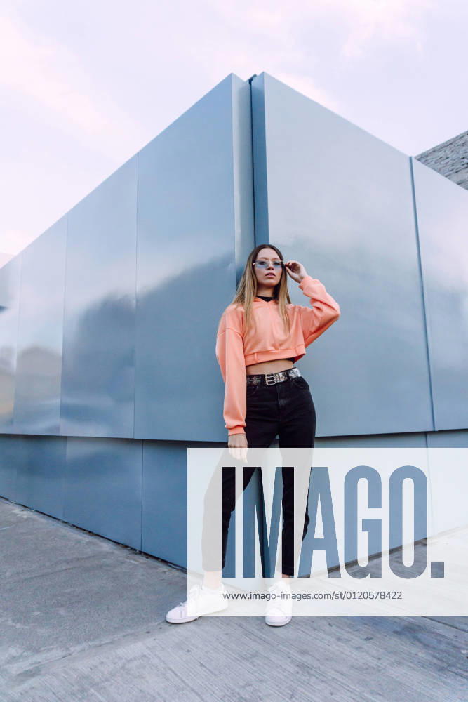 Confident female wearing jeans with top and orange shirt with stylish sunglasses standing near wall, Model released