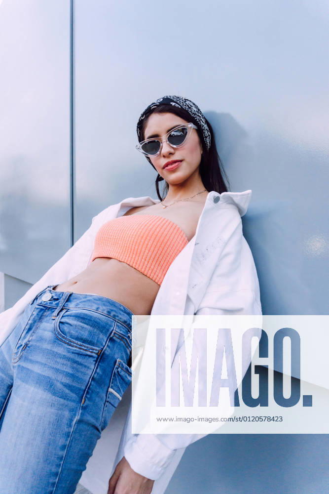Confident female wearing jeans with top and white shirt with stylish sunglasses standing near wall, Model released