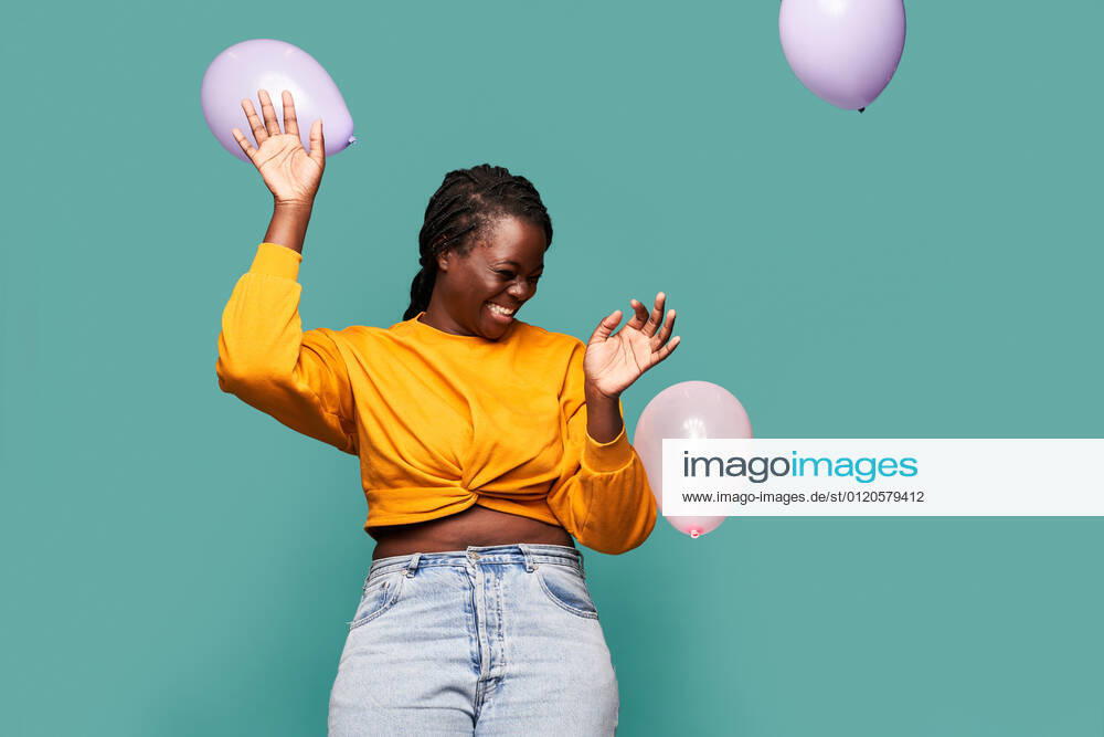 Excited African American female in jeans and yellow top standing near falling balloons against blue background in studio