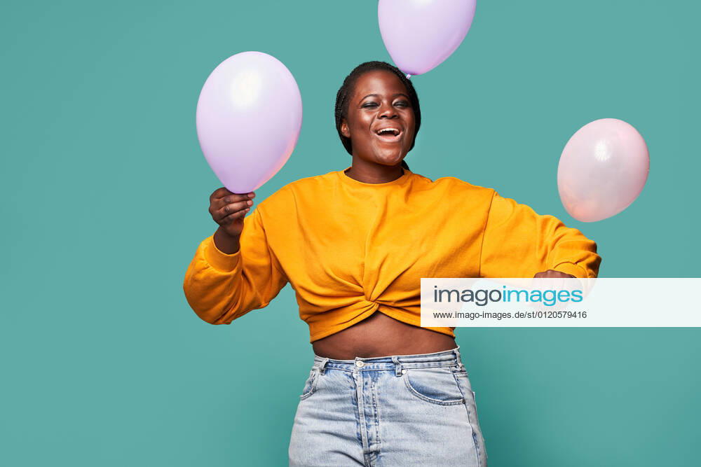 Excited African American female in jeans and yellow top standing near falling balloons against blue background in studio