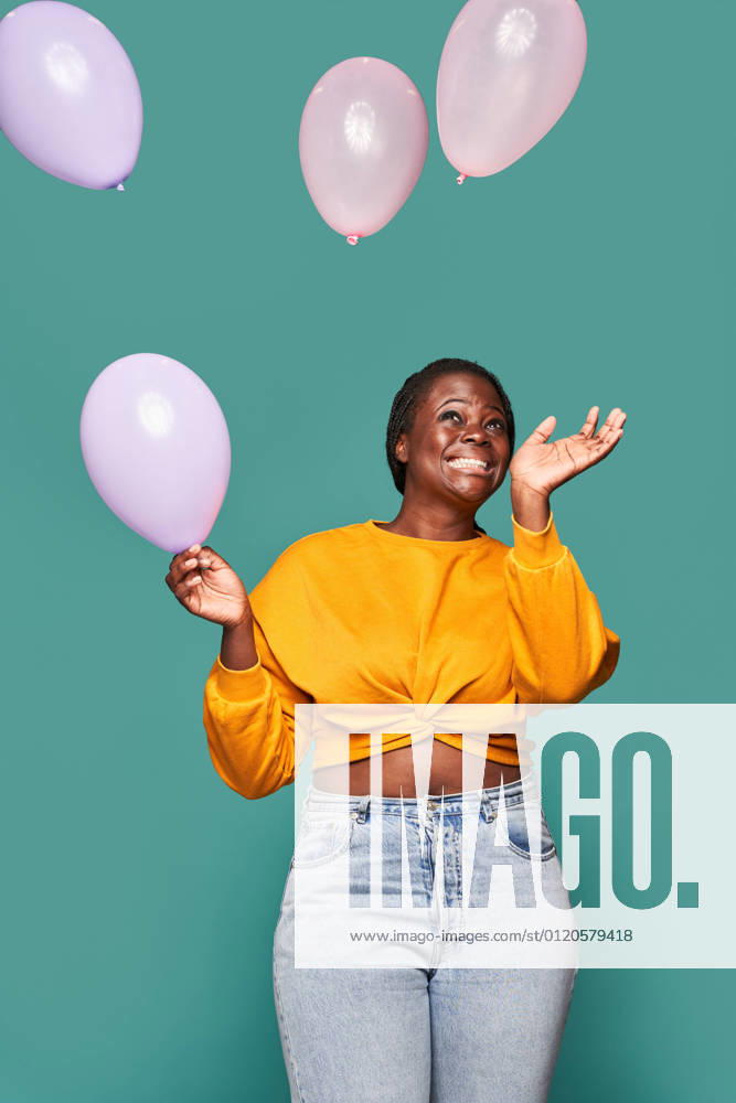 Excited African American female in jeans and yellow top standing near falling balloons against blue background in studio