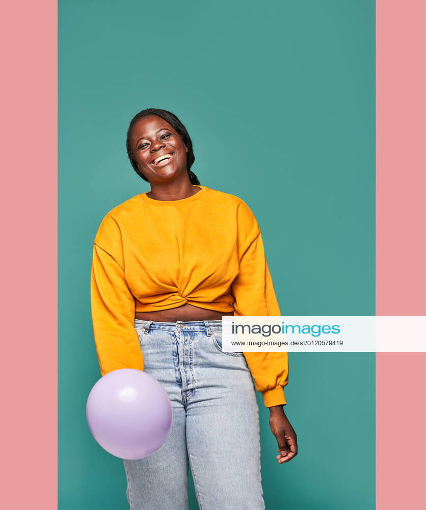 Excited African American female in jeans and yellow top standing near falling balloons against blue background in studio