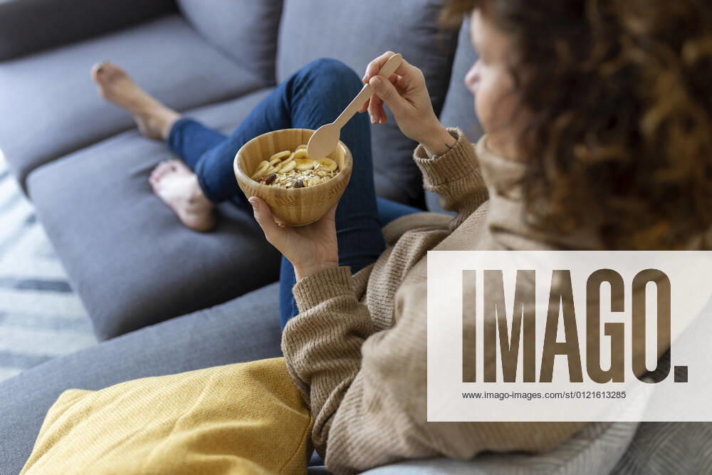 IMAGO Creative: Jahreszeit Winter, Couching Woman with food bowl in living room at home model released Symbolfoto property released JPTF00770