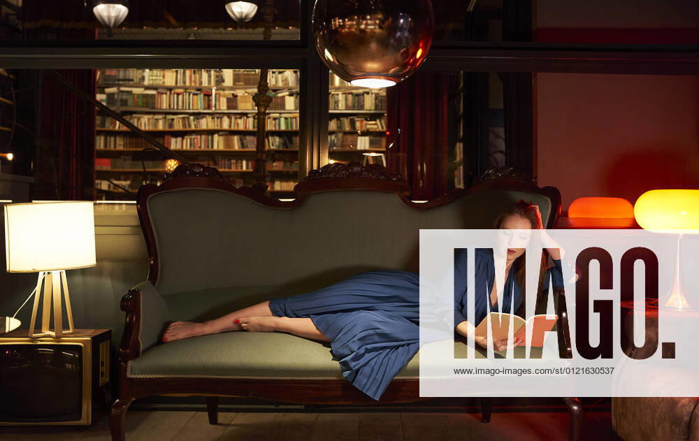 IMAGO Creative: Jahreszeit Winter, Couching Woman in blue dress reading book while lying on sofa at library model released Symbolfoto property released VEGF04352