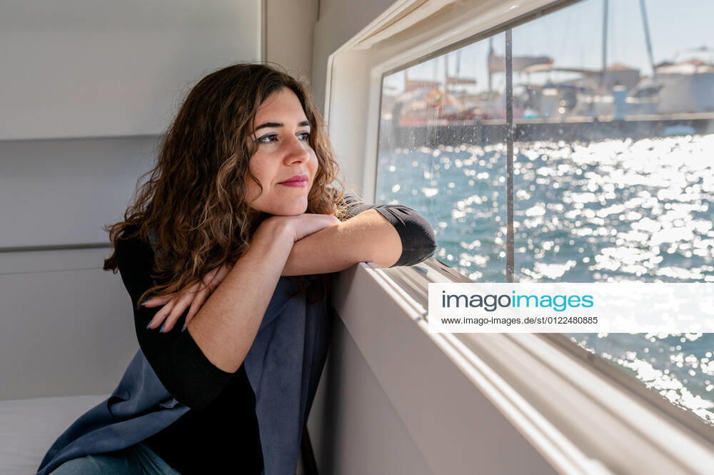 Tranquil female sitting in cabin of catamaran and looking out of window while admiring amazing view of sea on sunny day, Model released, Property released