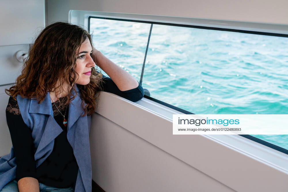Tranquil female sitting in cabin of catamaran and looking out of window while admiring amazing view of sea on sunny day, Model released, Property released