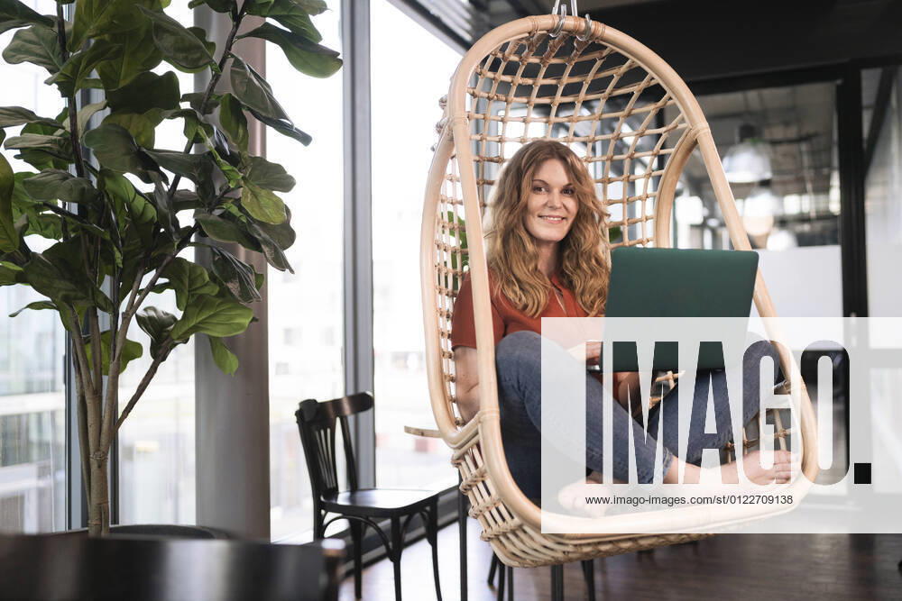 Smiling businesswoman with laptop sitting on swing chair at office cafeteria model released Symbolfoto property released FKF04337
