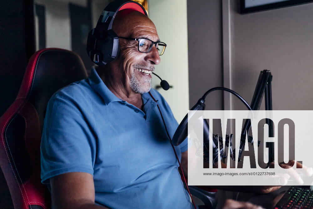 IMAGO Creative, Lifestyle, Gaming Smiling senior man wearing headphones using keyboard while playing video game at home model released Symbolfoto property released MEUF03131
