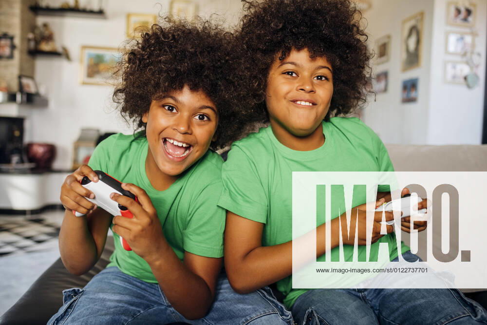 IMAGO Creative, Lifestyle, Gaming Cheerful twin boys with curly hair playing video games sitting on sofa in living room at home model released Symbolfoto property released MEUF03133