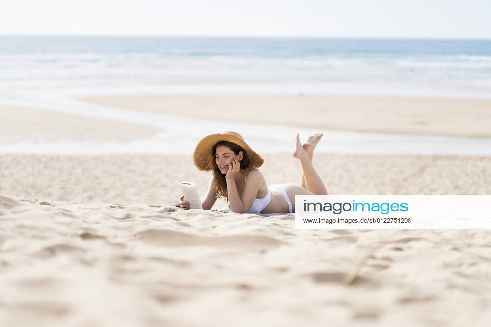 Young woman in bikini reading book while lying on sand during sunny day at beach model released Symbolfoto SBOF03852