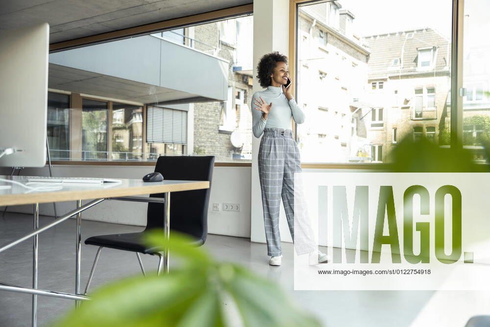 Smiling businesswoman gesturing while talking on mobile phone by window at office model released Symbolfoto property released UUF23265