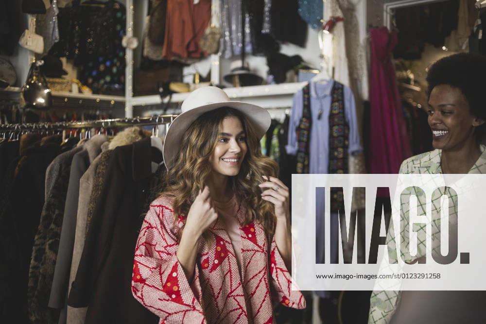 IMAGO Creative: Retro-Revival Smiling woman trying on hat while standing by female owner in clothing store Sweden, Stockholm, model released, property released